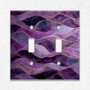 May include: A decorative double light switch cover with a watercolor design. The cover features a wavy pattern in shades of purple and gold. The design includes two toggle switch openings and two screw holes.