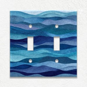 May include: A decorative double light switch cover with a blue wave design. The cover features a pattern of layered blue waves in various shades, creating a textured appearance. It has two switch openings and four screw holes.