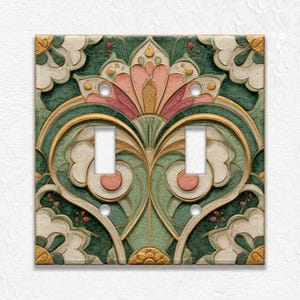 May include: Decorative double light switch cover with an Art Deco design. The cover features a symmetrical floral pattern in shades of green, pink, gold, and cream. The design is embossed, giving it a three-dimensional appearance. The cover has two switch openings and screw holes.