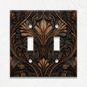 May include: A decorative double light switch cover with an Art Deco design. The cover features a detailed pattern in shades of brown, bronze, and black, with two white switch openings and four screw holes.