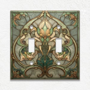 May include: A decorative double light switch cover with an Art Nouveau design. The cover features a floral pattern in shades of green, gold, and beige, with a stained-glass effect. The design includes two light switch openings and mounting holes.