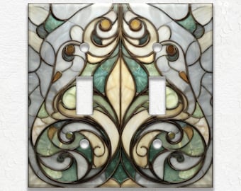 May include: A decorative double light switch cover with a stained glass design. The cover features a symmetrical pattern with swirls and floral elements in shades of green, cream, and brown, set against a light gray background.