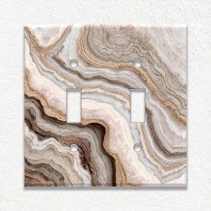 May include: A double light switch cover with a marbled design in shades of brown, beige, and white. The cover has two switch openings and two screw holes. The design resembles natural stone.