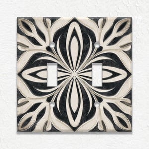 May include: A decorative double light switch cover with a black and cream floral pattern. The design features a symmetrical arrangement of stylized leaves and petals. The cover has two switch openings and four screw holes.