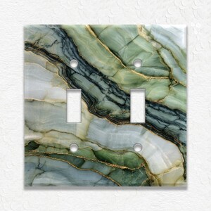 May include: A double light switch cover with a marbled design. The cover features shades of green, blue, and gold, resembling natural stone. The cover has two toggle switch openings and two screw holes.
