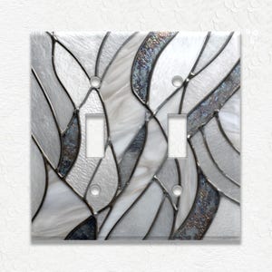May include: A decorative double light switch cover with a stained glass design. The cover features a pattern of white, gray, and iridescent blue-gray shapes, outlined with dark gray lines. The switch openings are rectangular.
