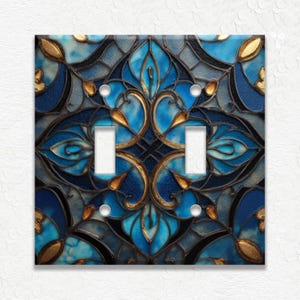 May include: A decorative double light switch cover with a stained glass design. The cover features a symmetrical pattern of blue, gold, and black shapes. The design includes floral and geometric elements, with two white light switch toggles.