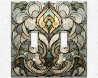 May include: A decorative double light switch cover with a stained glass design. The cover features a symmetrical pattern in shades of green, cream, gray, and gold, with a brown outline. The design includes floral and abstract shapes.