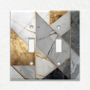 May include: A double light switch cover with a geometric design. The cover features a pattern of white, gray, gold, and black marble-like triangles, outlined with gold trim. The switch openings are rectangular.