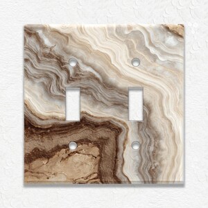 May include: A double light switch cover with a marbled design in shades of brown, beige, and cream. The cover has two rectangular openings for the switches and two small holes for screws.