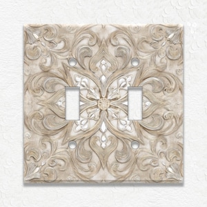 May include: A decorative light switch plate with a detailed, raised floral design. The plate features a neutral color palette of beige and off-white, with two switch openings and screw holes. The design is symmetrical and ornate.