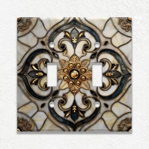 May include: Decorative double light switch cover with a detailed floral design. The cover features a cream-colored background with gold, black, and blue accents. The central design element is a gold floral medallion.