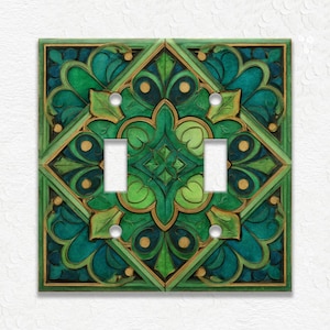 May include: A decorative double light switch cover with a carved design. The cover features a symmetrical pattern of green and teal leaves and geometric shapes, accented with gold. The light switch openings are white.