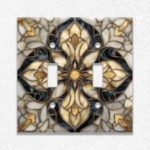 May include: Decorative double light switch cover with a stained glass design. The cover features a symmetrical floral pattern in cream, gold, and black, with a metallic gold outline. The switch openings are white, and the cover has a glossy finish.