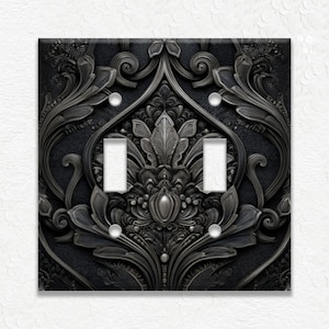 May include: A decorative double light switch cover with an ornate, embossed floral design in shades of gray and black. The cover features two toggle switch openings and four screw holes. The intricate pattern adds a touch of elegance to any room.