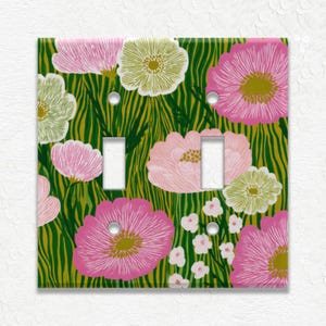 May include: A decorative double light switch cover featuring a floral design. The cover has a green background with yellow vertical stripes and pink and white flowers. The switch plate has two rectangular cutouts for the switches.