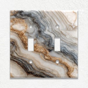 May include: A decorative double light switch cover with a marbled design. The cover features a swirling pattern of beige, gray, and brown tones, with two switch openings and two screw holes. The overall effect is a natural, stone-like appearance.