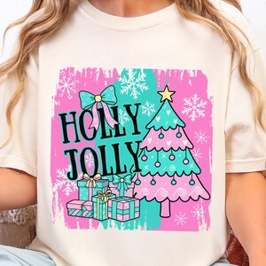May include: Cream-colored t-shirt featuring a colorful Christmas graphic. The design includes the words "HOLLY JOLLY" in black, a pink Christmas tree, presents, snowflakes, and a bow. The shirt is a festive holiday apparel item.