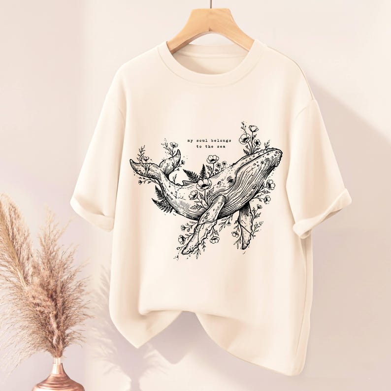 May include: Cream-colored sweatshirt with a black and white whale illustration surrounded by floral elements. The text "my soul belongs to the sea" is above the whale. The shirt is hanging on a wooden hanger.