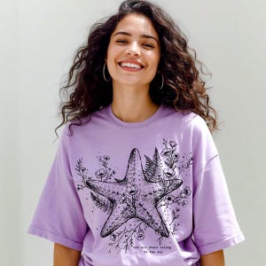 May include: A lilac-colored t-shirt featuring a detailed black and white starfish design, surrounded by floral and fern illustrations. The text "not all stars belong to the sky" is printed below the starfish. The shirt has short sleeves.