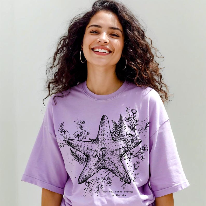 May include: A lilac-colored t-shirt featuring a detailed black and white starfish design, surrounded by floral and fern illustrations. The text "not all stars belong to the sky" is printed below the starfish. The shirt has short sleeves.