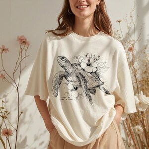 May include: Cream-colored t-shirt featuring a detailed black and white illustration of a sea turtle surrounded by flowers. The text "born of tide & moon" is below the turtle. The shirt has short sleeves and a relaxed fit.