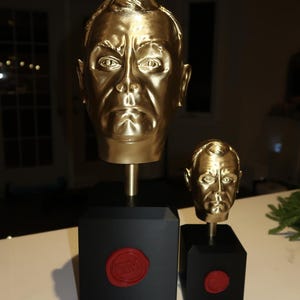 May include: Two gold-coloured busts of a man's head, one large and one small, mounted on black square bases. The bases have a red seal. The busts are detailed with facial features and sit on a white surface.