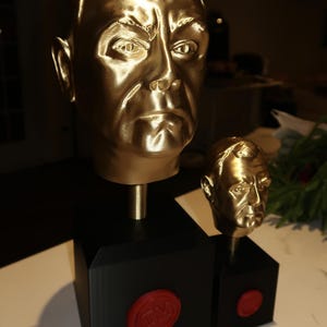 May include: Two gold-coloured busts of a man's head, one large and one small, are displayed. The busts are mounted on black square bases with a red seal. The larger bust is approximately 25 cm tall.