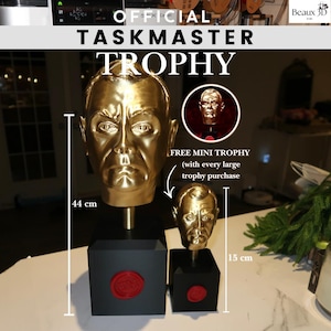 May include: A gold-colored Taskmaster trophy, 44 cm tall, with a bust of a man's face, mounted on a black base. A smaller, 15 cm tall, gold-colored mini trophy is also shown. The text "OFFICIAL TASKMASTER TROPHY" is displayed.