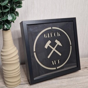 Könnte beinhalten: Schwarzer gerahmter Wandkunstdruck mit den Worten "GLÜCK AUF" und gekreuzten Hämmern in einem Kreis. Die Kunst wird auf einer Holzoberfläche mit einer beigen Vase und einer grünen Pflanze im Hintergrund präsentiert.