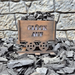 Könnte beinhalten: Ein hölzerner Wagen, gefüllt mit dunkler Holzkohle, vor einer Steinmauer. Der Wagen trägt die Worte "GLÜCK AUF" auf der Vorderseite. Der Wagen ruht auf einem Holzkohlebett.