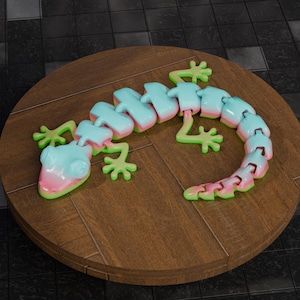 Peut inclure: Un jouet lézard articulé coloré dans les tons bleu clair, rose et vert. Le jouet est composé de segments connectés, permettant un mouvement flexible. Il repose sur une surface en bois ronde.