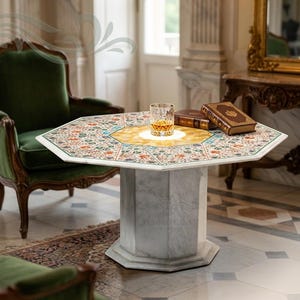 May include: An octagonal marble table with a decorative floral pattern top. The table features a central gold-colored circle and a white marble base. A glass of amber liquid and two books are on the table. The table is in a room with green chairs.