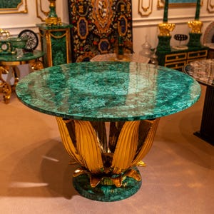 Puede incluir: Una mesa redonda con una tapa de malaquita verde y una base dorada en forma de pétalo. La mesa es una pieza llamativa, que muestra lujo y un diseño intrincado. El diámetro de la mesa es de aproximadamente 122 cm.