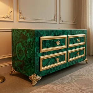 Puede incluir: Cómoda verde y negra con estampado de malaquita y cuatro cajones. Cada cajón está enmarcado en dorado con tiradores dorados ornamentados. La cómoda tiene patas doradas decorativas y está sobre una pared neutra.