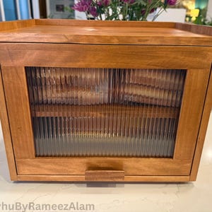 May include: A wooden bread box with a light brown finish. The box has a rectangular shape with a glass panel door featuring vertical ribbed detailing. A small wooden handle is centered on the door. The top of the box is flat.