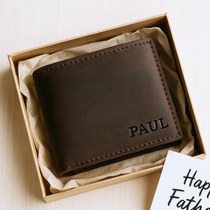 Puede incluir: Una cartera de cuero marrón con el nombre "PAUL" grabado en la parte delantera, presentada en una caja de regalo. Una tarjeta blanca con "Happy Father's Day" en escritura negra está al lado de la caja. La cartera es de estilo plegable.