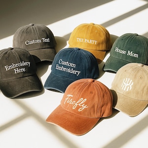 Puede incluir: Una colección de gorras de béisbol en varios colores, incluyendo gris, amarillo mostaza, azul marino y naranja óxido. Cada gorra presenta texto bordado como "Custom Text", "The Party", "House Mom", "Custom Embroidery" y "Firefly".
