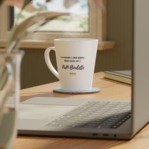 May include: A white ceramic latte mug with a curved handle. The mug features black text that reads "Le monde a des papis... Mais nous, on a Papi Boulette" with three small gold hearts. The mug sits on a blue coaster.
