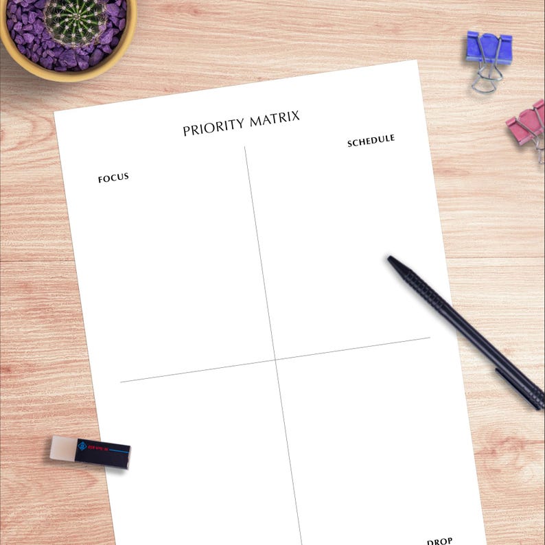 Pode incluir: Uma folha de papel branca com as palavras "PRIORITY MATRIX", "FOCUS", "SCHEDULE" e "DROP" impressas. Uma caneta preta, clipes azuis e rosa e uma borracha est&atilde;o sobre uma superf&iacute;cie de madeira. Um pequeno vaso com um cacto est&aacute; no canto superior esquerdo.