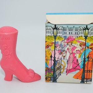 Op de afbeelding: Een roze Avon laarzenvormig verzamelobject naast een doos met een vintage illustratie van mensen in een park. De laars heeft bloemendetails en het Avon-logo. De doos toont een kleurrijke scène met figuren in historische kleding.