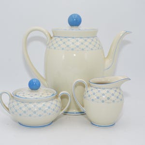 Könnte beinhalten: Cremefarbenes Teeservice mit blauem und weißem geometrischen Muster. Das Set beinhaltet eine Teekanne, eine Zuckerdose mit Deckel und einen Sahnekännchen. Jedes Teil hat einen blauen, kugelförmigen Knopf und blaue Verzierungen.