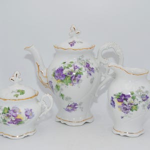 Op de afbeelding: Een wit porseleinen theeservies met een theepot, suikerpot en melkkan. Elk stuk is versierd met een bloemmotief van paarse en witte bloemen, geaccentueerd met gouden randen. De set is ontworpen voor het serveren van thee.