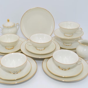 Könnte beinhalten: Ein cremefarbenes Teeservice mit goldenen Verzierungen. Das Set umfasst Teetassen, Untertassen, Teller, eine Zuckerdose und einen Sahnekännchen. Die Tassen und Untertassen sind gestapelt. Die Teller sind rund.