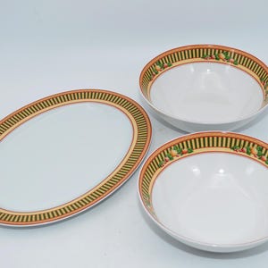 Könnte beinhalten: Ein Set aus weißen Keramik-Servierschalen. Das Set besteht aus zwei runden Schalen und einer ovalen Platte. Jedes Stück hat einen dekorativen Rand mit einem gestreiften grün-gelben Muster, akzentuiert mit roten und grünen Fruchtmotiven.