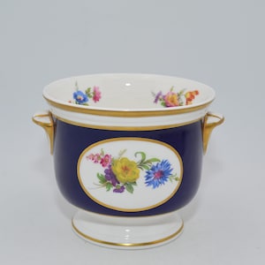 Op de afbeelding: Een porseleinen plantenbak met een marineblauwe buitenkant, witte binnenkant en gouden rand. De plantenbak heeft een ovaal bloemmotief met gele, blauwe en paarse bloemen. Twee gouden handvatten bevinden zich aan weerszijden.