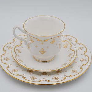 Peut inclure: Service à thé blanc avec des détails floraux dorés. La tasse a une anse incurvée et des bordures dorées. La soucoupe et l'assiette présentent un motif floral doré assorti et des bordures dorées. L'ensemble est sur fond blanc.