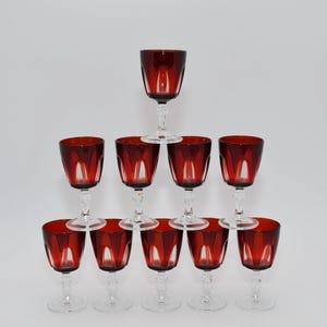 Op de afbeelding: Een set van tien glazen op voet, gerangschikt in een piramidevorm. Elk glas heeft een dieprode kelk met een heldere glazen steel en voet. De kelken hebben een opvallend verticaal paneelontwerp en de heldere stelen zijn gefacetteerd.