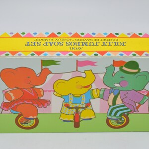 Op de afbeelding: Een vintage Avon Jolly Jumbo Soap Set doos. De doos bevat illustraties van drie cartoon olifanten die op een eenwieler rijden. De olifanten zijn in oranje, geel en groen, met een circus-thema. De doos heeft een gele banner met tekst.