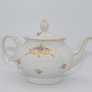 Può includere: Teiera in porcellana bianca con coperchio, manico e beccuccio. La teiera è decorata con finiture dorate e disegni floreali in rosa, giallo e blu. Ha un corpo bombato e poggia su una piccola base.